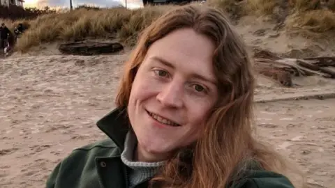 Ryan Castellucci A person with long hair smiles at the camera taking a selfie. They're sat on a beach wearing a green coat and a white roll-neck jumper.