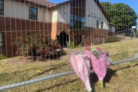 Two floral bouquets in pink wrapping, laid against metal fencing that surrounds the crash site. The damaged wall is in the background across a grassed area.
