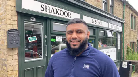 A bearded man in a blue fleece top standing in front of his shop.