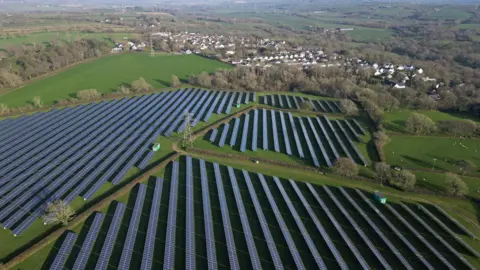 A drone shot ona clear day showing solar panels stretching across a large area of fields, with blocks of various shapes bisected by hedgerows and then green fields surrounding the development with wooded areas and the village with its white stucco homes and brown roofs, some close to one end of the solar park, but with a signficant wooded area in between . 