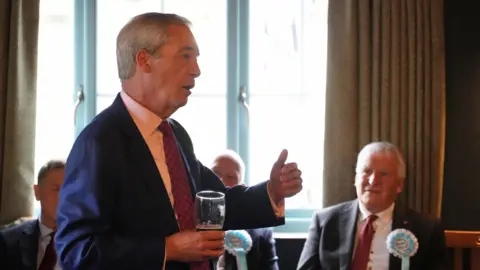 Nigel Farage is in a pub talking to a group of people wearing blue and white rosettes. He is holding a pint glass and three men are seen listening to him.