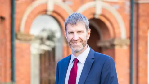 Handout A man with a beard smiling at the camera. He is in front of a red brick building and is wearing a suit and tie