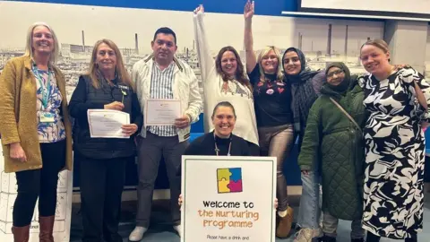 Seven women and a man stand with their arms around each other, smiling. Another woman crouches in front of the group holding a sign that says 'Welcome to the Nurturing Programme'. 