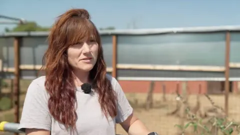 Lucie Francis stands in front of the bear enclosure. She has long, wavy red hair with a fringe and wears a light grey short sleeved cotton t shirt. She looks slightly off camera while giving an interview.