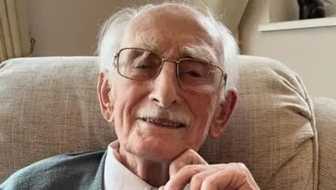 Man wearing glasses looks straight into the camera lens. He's smiling and wearing a white shirt with a blue and white spotted tie and grey cardigan