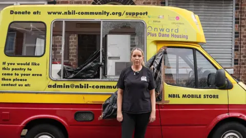 Michelle Roach stands in front of the damaged van. She has long, blonde hair pulled back from her face. She wears a black T-shirt, black trousers and black and white trainers. The van is painted yellow and red. The company details are written in black font on the sides. The side windows have been smashed in, a bin bag is visible inside.