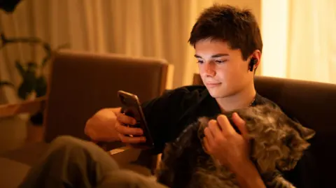 Getty Images A teenage boy sits on the sofa looking at his smartphone. He has earphones in and short dark hair. He is holding a cat.