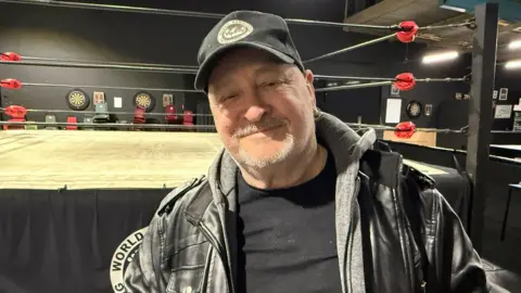 Rob Butler/BBC Wrestler Ricky Knight stood beside a ring smiling. He has a grey goatie beard and is wearing a black jacket and baseball cap