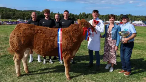 CJS PHOTOGRAPHY Lee Cain, holding the trophy, stands with his family and helpers alongside a large brindle coloured cow with a ribbon over its shoulders.