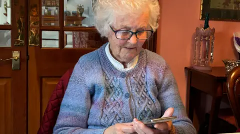 Vera is an elderly woman, with short white fluffy hair. She is sitting on a chair and looking down at her smartphone in her hands. She is wearing a blue and purple knitted cardigan and black glasses. 