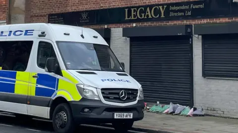 A police van parked in front of a red and grey brick building. A black sign on the front of the building reads: Legacy Independent Funeral Directors Ltd. On the ground are a dozen bouquet of lowers laid in a row against black shutters.