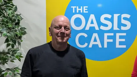 Colin McGregor-Paterson smiles directly at the camera and is wearing a black T-shirt. He is standing in front of a blue and yellow sign which reads "The Oasis Cafe".