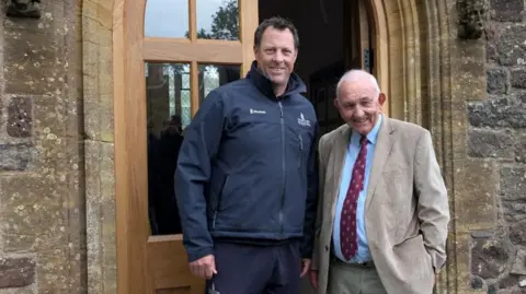 Marcus Trescothick and Richard Walsh stand next to each other in front of the church door. They are both smiling at the camera.