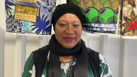 Elizabeth Baines / BBC A woman wears a black headscarf and green dress. Behind her, traditional African fabrics hang.