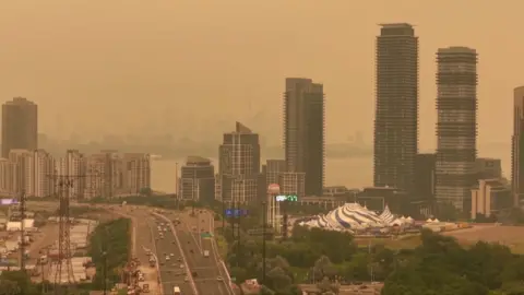Orange haze fills the sky over a skyline of tall buildings and a highway in Toronto.