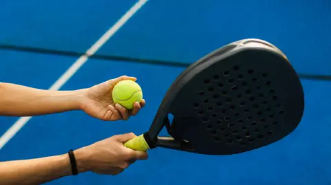 A Padel ball is held in one hand and a black Padel racket in the other. The court is blue with white lines and a black net. The person has a brown band around their right wrist. 