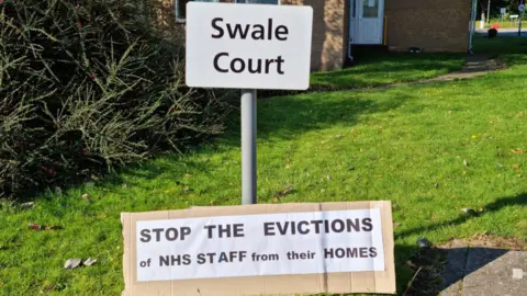 Supplied A white post in the grass outside a red brick building reads 'Swale Court'. On the ground is a handmade cardboard sign which reads: "Stop the evictions of NHS staff from their homes."