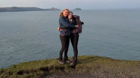 Handout Tara and Sophia hug each other on a cliff overlooking the beach, there is a stretch of sea behind them and a small island in the ocean