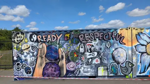 Nue A mural which has different images, including a finger and thumb holding two purple rocks, a space ship and space scene, a bird and a large flower which has yellow centre and white flowers.