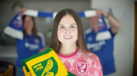 Jamie Niblock/ BBC Eloise King has brown hair, and stands in front of her mother and father. She is wearing a pink Norwich City kit, and has a yellow and Green Norwich City scarf over her shoulder. Tracey King (left) and Chris King stand in the background wearing blue Ipswich Town shirts. They are holding a blue and white scarf between them.
