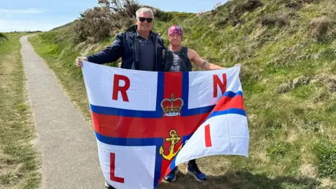 RNLI Geoff Rice (right) stands with Stuart Blackley, a taller man wearing sunglasses as they hold the RNLI flag on Peel Hill.