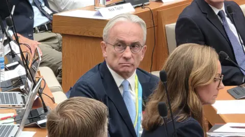 Paul Moseley/BBC David Bick wearing a dark blue suit jacket, white shirt and light blue tie at a council meeting. He is surrounded by a group of people seated at desks in a surrounded by microphones, laptops and documents.