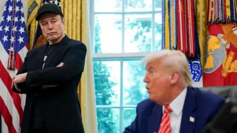 U.S. President Donald Trump and Elon Musk attend a press conference in the Oval Office of the White House. Musk is glancing sideways at Trump, who is seated and speaking, while slightly out of focus. 