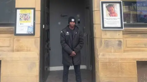 Believe Achieve CIC Tunde Olasupo standing in the doorway of Believe Achieve CIC, wearing a black coat and had. 