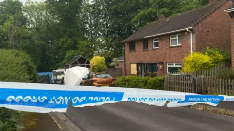 BBC Blue police tape is seen in front of a house with boarded up windows and a white forensics tent.