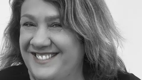 The Kelly Foundation A close-up black and white photo of Sarah Forrester's face. She is smiling at the camera and has glittery eyeshadow on and shoulder length hair.