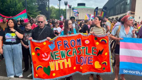 Robin Davies/BBC A group of people standing in front of Newcastle's Civic Centre. Three people at the front are holding a handmade banner which reads 'From Newcastle with love' It is red and has rainbows, hearts and lightening bolts stitched onto it. The people holding it are looking down and the people around them are waiting for the march to start. To the right, the corner of a blue, pink and white transgender pride flag is visible.