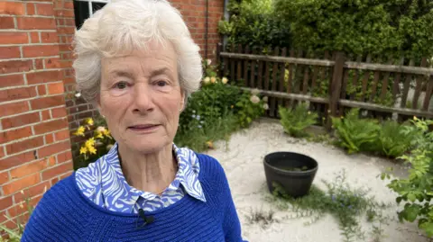 A woman with white hair looks into camera with her damaged garden behind her