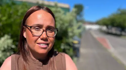 Ms Gracey has long brown hair, is wearing makeup and has brown round glasses on. She is smiling at the camera as she wears a brown buttoned top.
