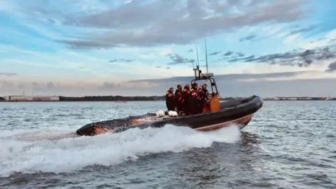 Hamble Lifeboat A lifeboat crew stand together on a boat, which looks to be moving at sea. They are all wearing red reflective clothing.