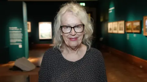 Qays Najm/BBC A woman standing in the middle of an art exhibition. She has shoulder length grey hair and has glasses as well as a black top with some white specks on it. 