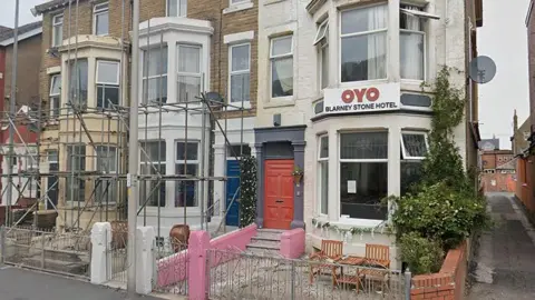 A white guesthouse on the corner of a street with a sign at the front which reads OYO Blarney Stone Hotel. Other properties to the left of the street have scaffolding up.