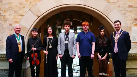 Bristol Beacon Six young men and women stand beneath a stone archway with two older men, one of whom is a Lord Mayor with a chain of office round his neck. The young people are looking to camera and one is holding a violin and another has a flute. Two of the young musicians are wearing medals