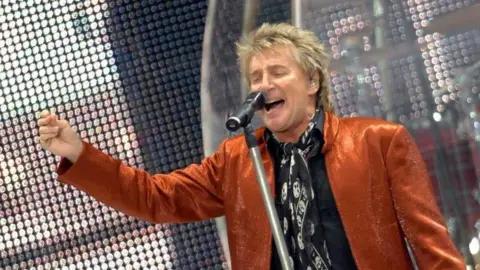 Ashley Pickering Rod Stewart performs at Portman Road on stage. He is wearing an orange suit jacket and a black shirt. He has one arm raised as he sings into a microphone.