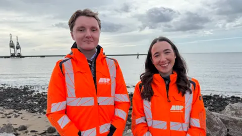 Two people in hi-vis orange jackets. On the left is a man with short brown hair and on the right a woman with shoulder-length brown hair, who is smiling at the camera. Behind them is the Celtic Sea.