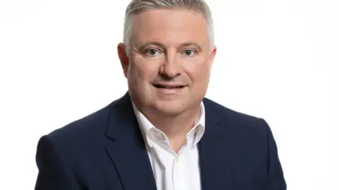 Mid and South Essex ICB A head and shoulders shot of Paul Scott against a white background. He is wearing a navy suit jacket and white shirt. He has grey hair and is smiling.