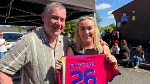 A man, with short grey hair, wearing a short sleeve green shirt, with his hand around a woman, with long blonde hair. The woman is holding up a pink jersey with 'Fleadh 26' written on the back. 