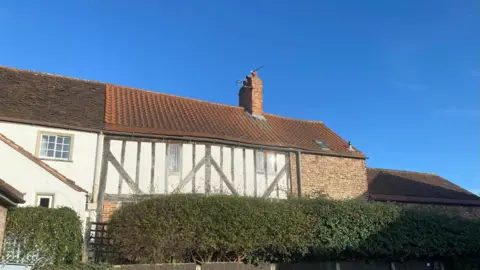 A shot of the rooftops of a row of terrace houses, over a hedgerow. The middle house is a half-timber building, with a red roof. 