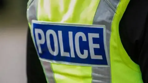 A person wearing a black long sleeved top and a yellow hi-vis jacket, with grey stripes and a blue patch in the middle with the wording 'police' in white lettering.