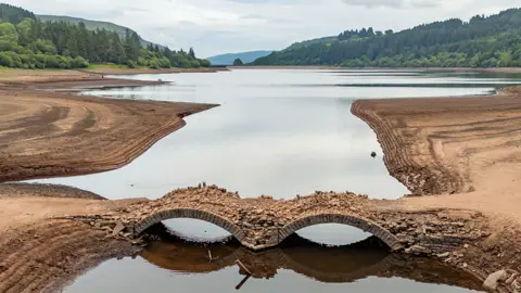 Two-arch stone bridge emerges in a largely dry bed forming part of today's reservoir