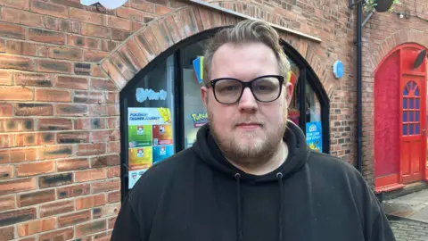 Photograph of Aaron Manders, who runs the Evolution Trading Cards store in Jaxon's Court. The image of the 34 year-old is taken outside of his shop. 