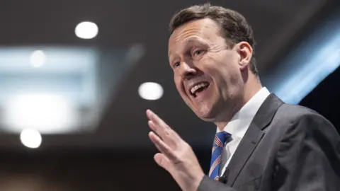 PA Media A man with combed back dark hair speaks with his left hand in front of him. He is wearing a dark suit, white shirt and blue and red tie.