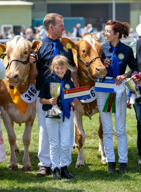 James Warren Three people standing with award winning light brown Guernsey cows, a girl aged 9 in the middle of them holding a trophy