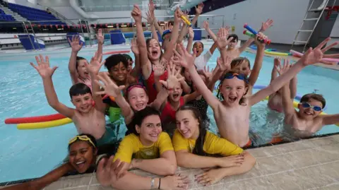 A group of smiling children in a pool with their arms outstretched. There are red and yellow noodles in the water and some of the children are wearing googles. 