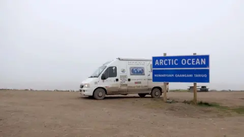 Tread the Globe A white camper van parked on a barren dirt patch in front of a blue sign that says Arctic Ocean
