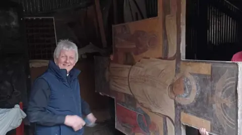 Reverend Sheila Bawden Vicar smiling at the camera while the painting is carried on its side out of the barn 